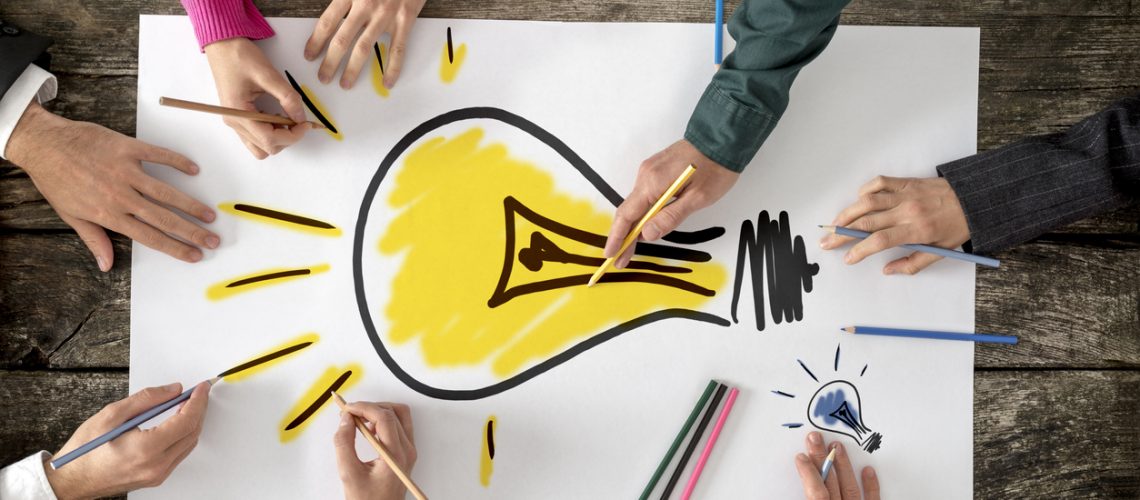Top view of six people, men and women, drawing bright yellow light bulb on a large sheet of paper or placard. Conceptual of teamwork, research, education and innovation.