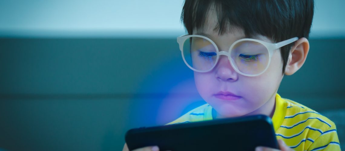 Cute little child watching cartoon on smartphone in the dark room. Dangers of blue light damage his eyes. Handsome little boy can get age related macular degeneration of blue light, he wear eyeglasses