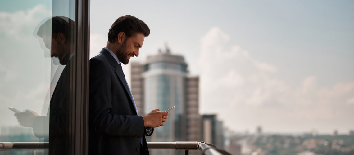 Take a pause. Waist up portrait of smiling businessman reading massages on smartphone while standing on office terrace. Copy space on right
