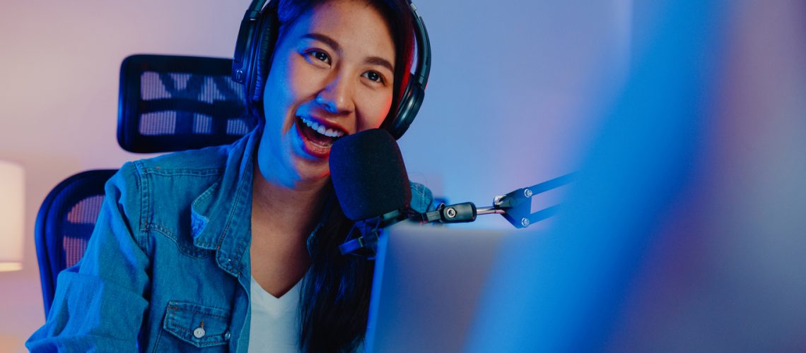 Happy asia girl host record podcast use microphone wear headphone with laptop interview guest conversation for content in her home studio at night. Sound equipment concept. Content creator concept.