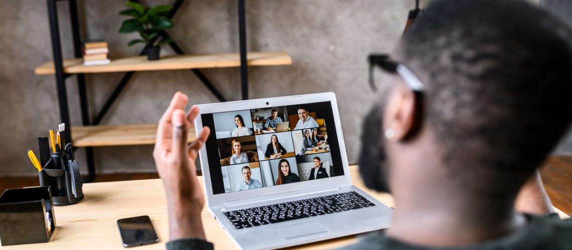 Confident African-American male worker talking online with coworkers, back view of black guy speaks and gestures to many people on video screen. Remote work, virtual meeting